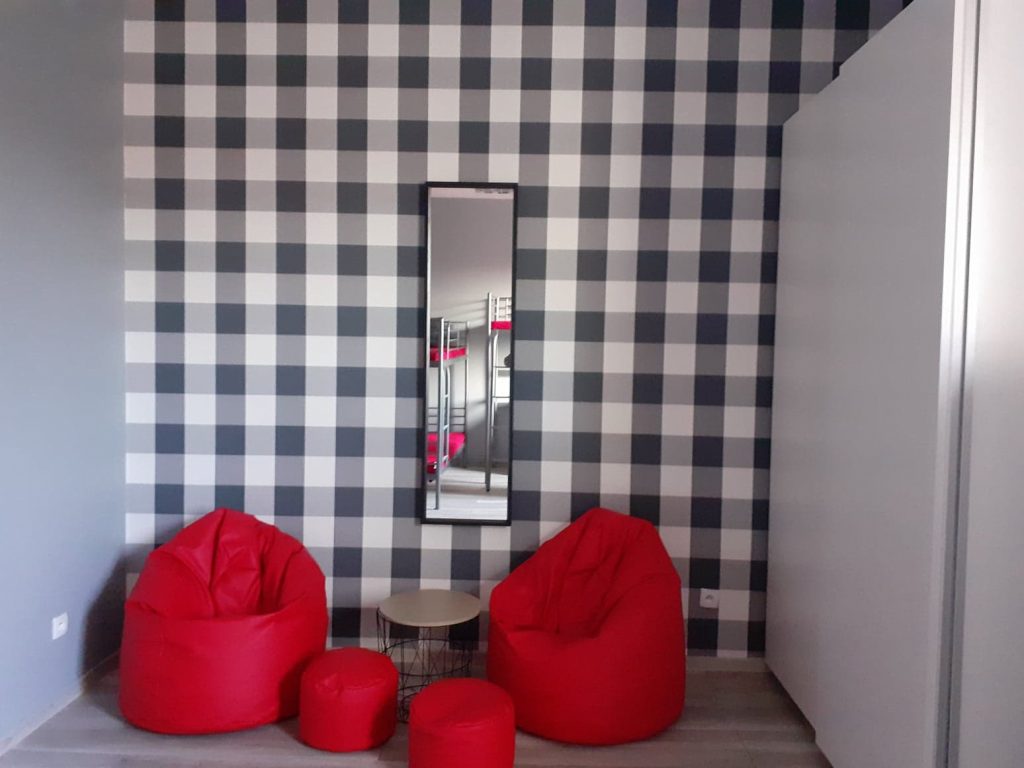 tapeta w czarno-białą kratę, na jej tle stoją czerwone pufy i stolik na ścianie wisi czarne lustro